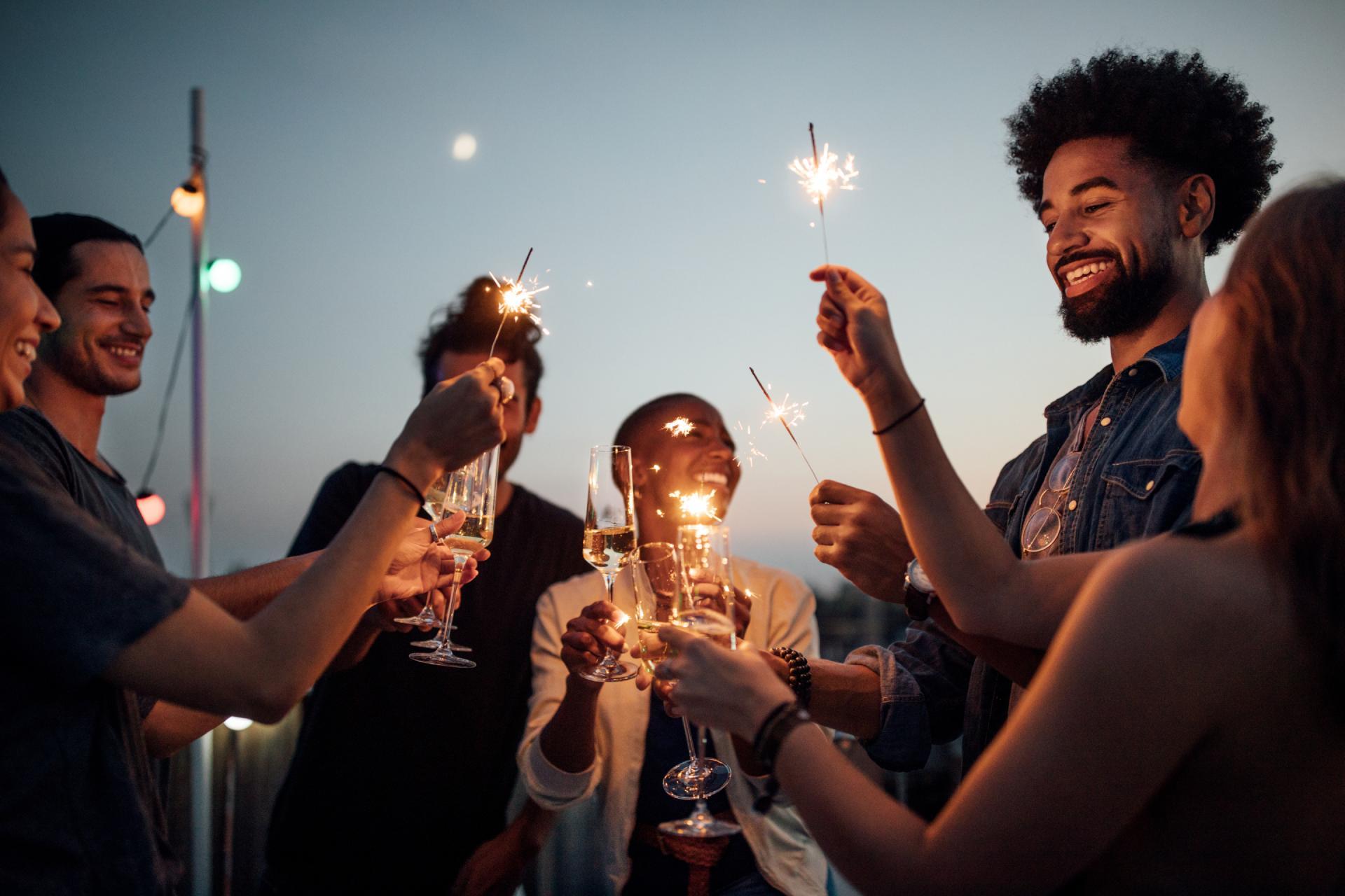 A multiethnic group of people toasting champagne glasses and holding sparklers at twilight