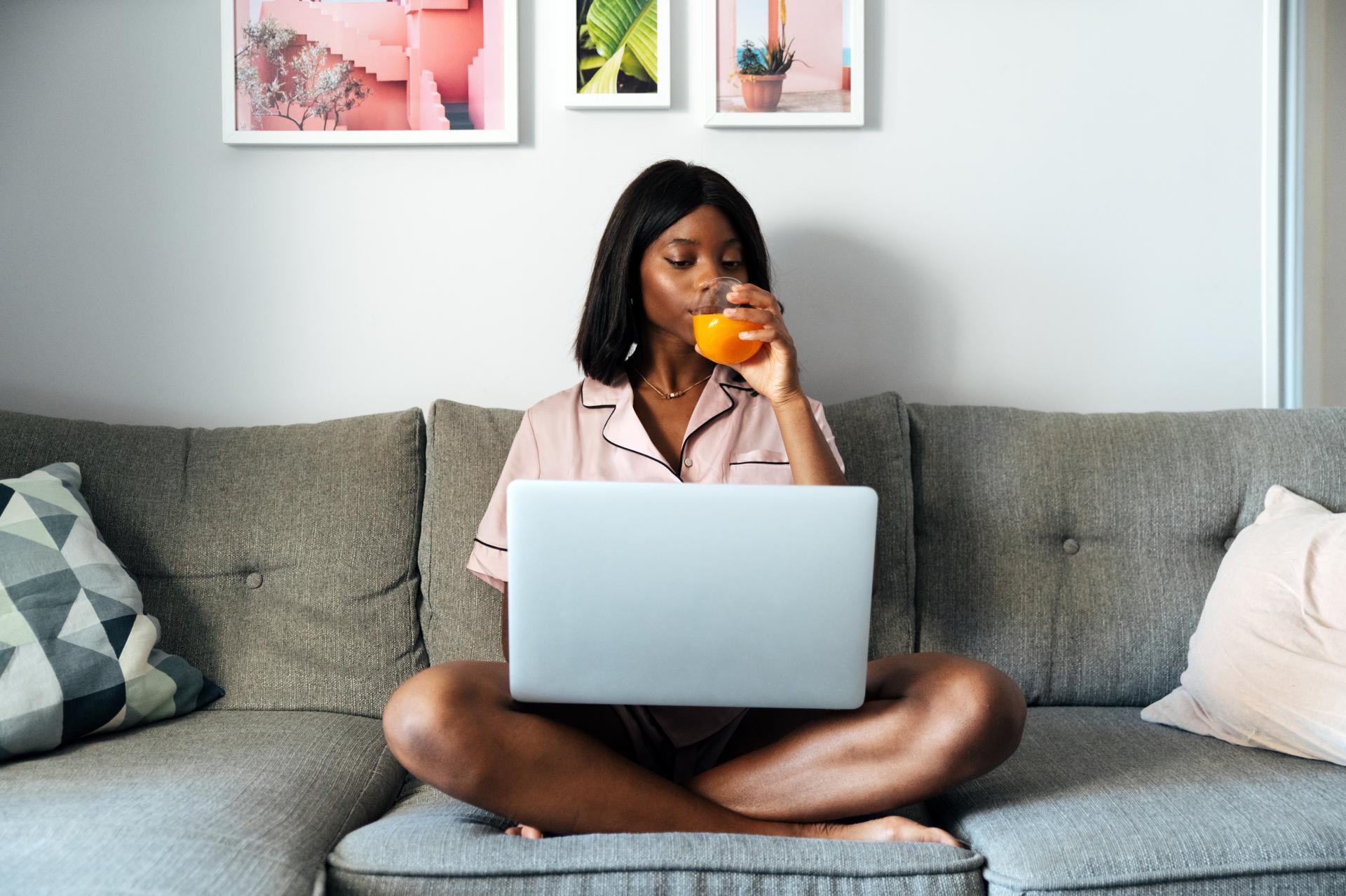 A Black woman in pajamas sitting cross-legged on the couch drinking orange juice as she looks at her laptop