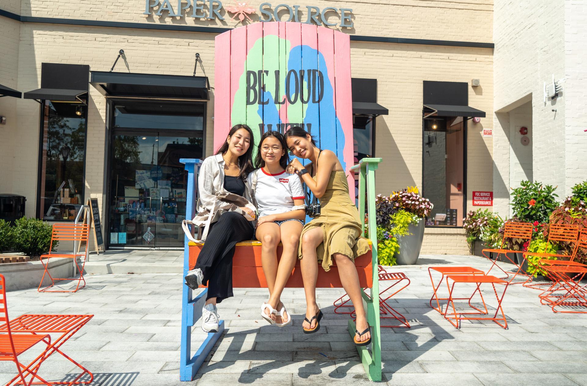 Three young Asian women sitting on a giant Pride chair in front of Paper Source at Suburban Square