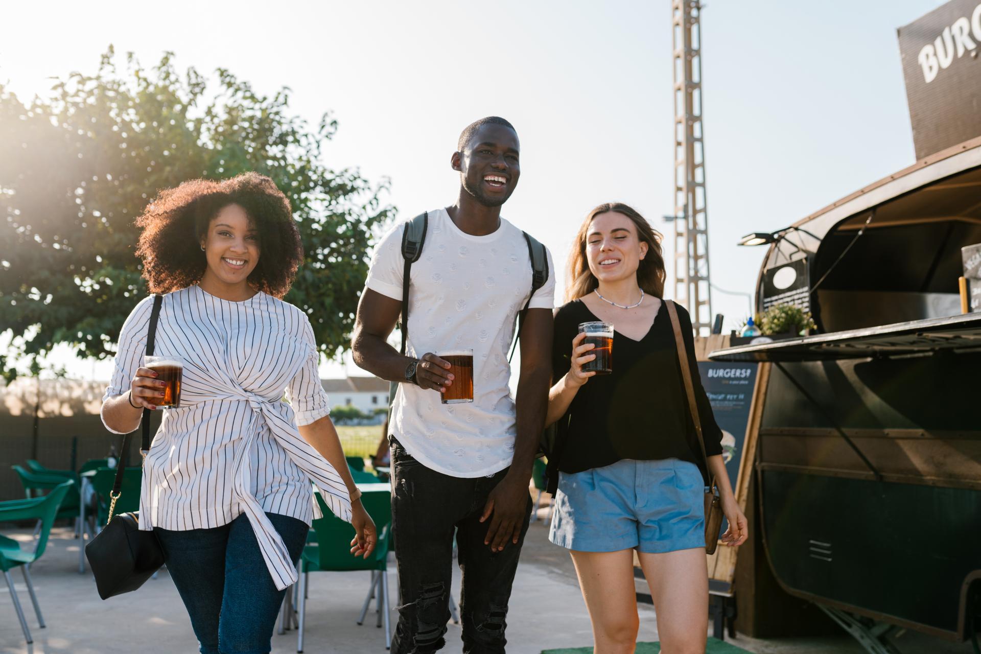 A group of young adults carrying beers and talking animatedly as they walk past a food truck